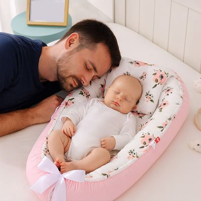 Un bébé qui dort dans un réducteur de lit pour bébé avec son père à côté.